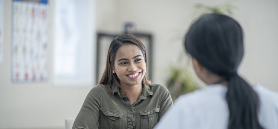 A young woman of Indian ethnicity smiles while discussing her health concerns with her doctor. The doctor has her back towards the camera and is wearing a white scrub shirt.
