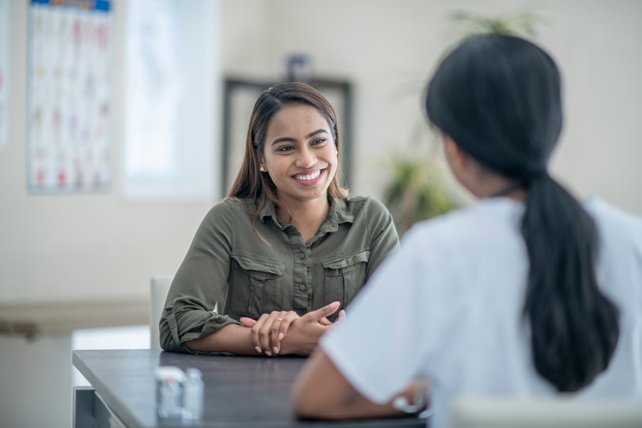 A young woman of Indian ethnicity smiles while discussing her health concerns with her doctor. The doctor has her back towards the camera and is wearing a white scrub shirt.