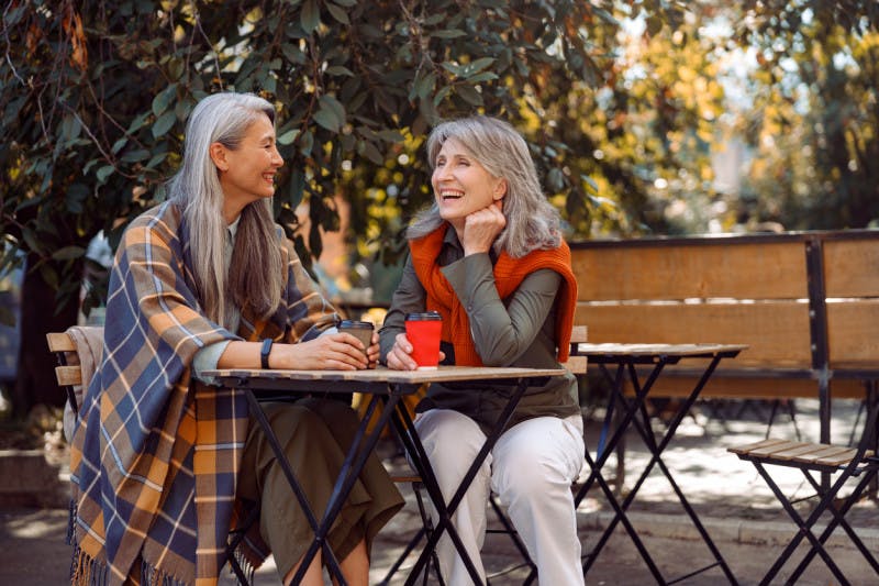 Mature Asian lady with positive grey haired friend spend time together sitting at small table in street cafe on nice autumn day