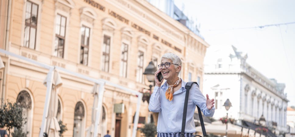 Portrait of Senior Woman with Grey Short Hair in the City District. Older Business Woman with Eyeglasses is Carrying a Briefcase and Talking over her Mobile Phone During the Walk in the City Center.