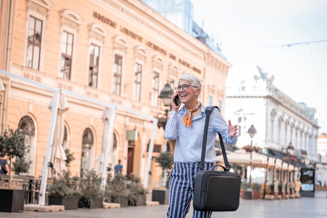 Portrait of Senior Woman with Grey Short Hair in the City District. Older Business Woman with Eyeglasses is Carrying a Briefcase and Talking over her Mobile Phone During the Walk in the City Center.