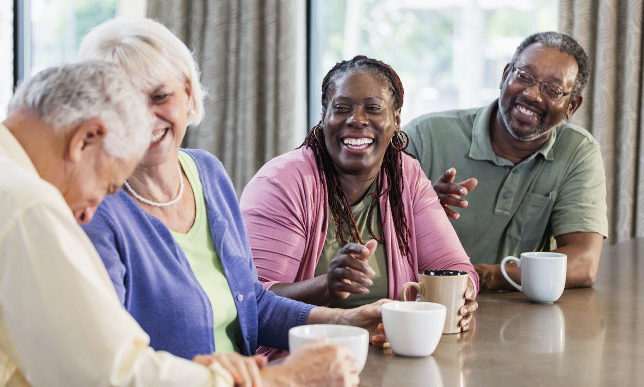 Four multi-ethnic mature and senior adults sitting indoors at a table, drinking coffee, conversing and smiling. The main focus is on an African-American couple in their 50s. Their friends are a senior couple in their 70s.