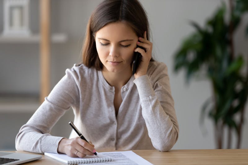 Pretty focused businesswoman sitting at desk holding cellular talking with client holding pen note on notepad