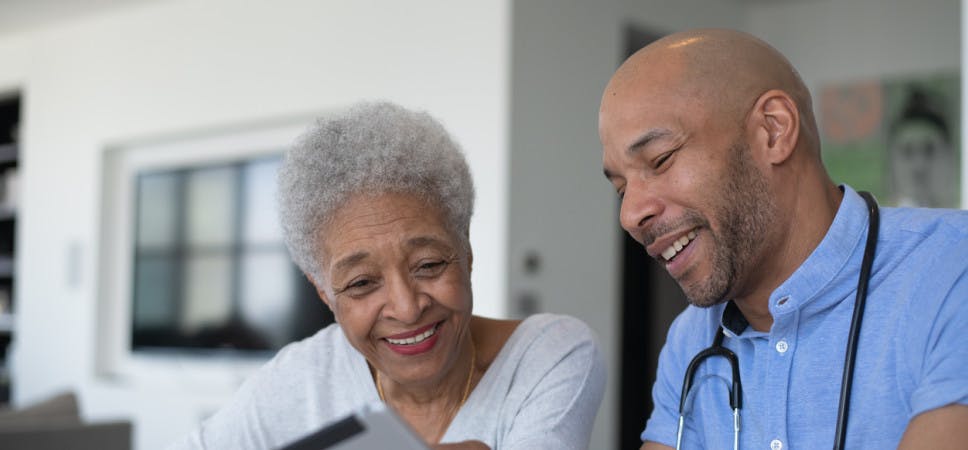 A young mixed race male doctor meets with his senior African American patient in a casual setting. They are seated at a table as they talk and both are wearing casual clothing. The doctor has a stethoscope around his neck and is holding out a tablet as they talk about the results. Both are smiling as they discuss the good news.