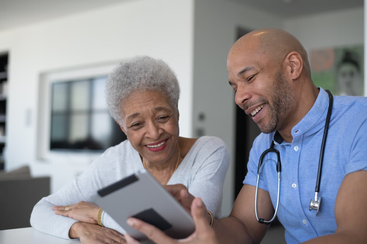 A young mixed race male doctor meets with his senior African American patient in a casual setting. They are seated at a table as they talk and both are wearing casual clothing. The doctor has a stethoscope around his neck and is holding out a tablet as they talk about the results. Both are smiling as they discuss the good news.