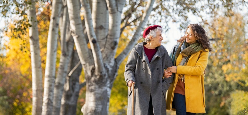 Granddaughter walking with senior woman in park wearing winter clothing. Old grandmother with walking cane walking with lovely caregiver girl in sunny day. Happy woman and smiling grandma walking in autumn park.
